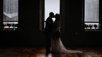 Couple standing side- by- side. holding hands in an apple orchard. Bride is wearing a low cut white dress with lace accents. She has a green and whire bouquet in her right hand and has a cathedral length veil. Groom is wearing a grey suit with a white tie and is standing with his left hand in his pocket