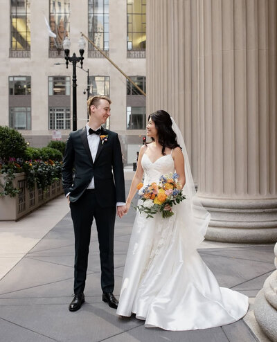 Brunette bride with classic gown and vibrant bouquet smiles in front of large pillar outside the venue while holding hands with groom wearing a black tux.