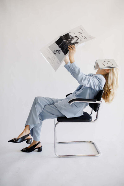 A woman sitting in a chair with a book on her face holding a newspaper posing for a photo