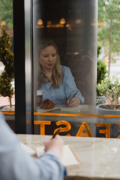 Photo taken through a cafe window of Anna Bradshaw writing copy at a table