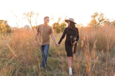 A couple walks hand in hand through a grassy field at sunset, with the pregnant woman holding her belly and wearing a wide-brimmed hat.