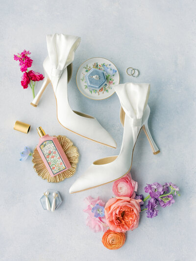 White satin pointed toe heels lay on blue flat lay mat with bow on sides and colorful pink, purple, and orange flowers and bride's jewelry and perfume on ornate dish