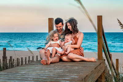 A family of a mom, dad, and two toddlers are sitting on a wooden path leading to the beach behind them.