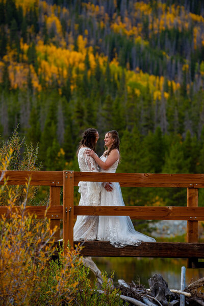 Two brides standing on a wooden bridge with  yellow trees surrounding them.