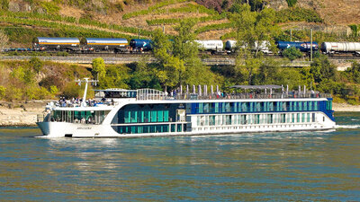 A modern white river cruise ship with large glass windows sails along a wide river, with green vineyards and a passing freight train visible on the hillside behind it.