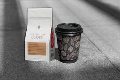 A white coffee bag labeled "Morning Oak Coffee, Heritage Oak Blend" with a gold accent sits on a light gray background. Beside it, there’s a black coffee cup with an oak leaf pattern, a black lid, and a smooth, matte finish.