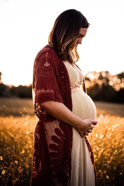 Femme enceinte dans un champ, mains sur le ventre au soleil couchant