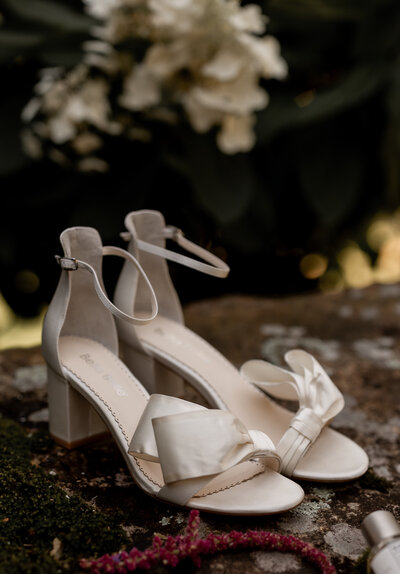 A pair of elegant white high-heeled sandals with ankle straps and large bows is placed outdoors on a stone surface. In the background, blurred white flowers and green foliage are visible.