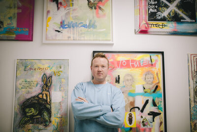A man in a light blue sweatshirt stands with arms crossed in front of a gallery wall filled with colorful, expressive mixed media paintings featuring abstract figures, animals, and text elements.
