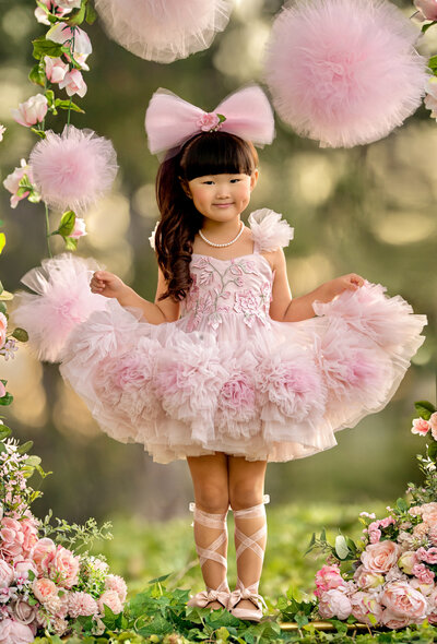 Asian girl in pink tulle tutu dress with pom poms standing in a field of low brush surrounded by pink flowers and pink tulle pom poms with a large pink bow in her hair and sheer ribbons laced up her shoes on both legs.