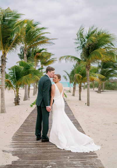 a bride with a long white veil and groom in black tuxedo kiss in front of a white barn