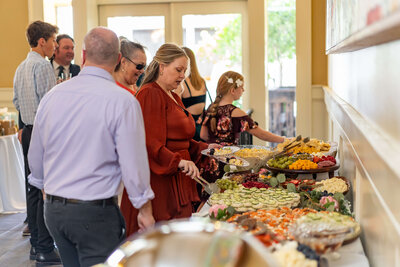 People dressed in semi-formal attire serve themselves food from a buffet table with a variety of dishes, including fruit, crackers, dip, and hot entrees, at an indoor social event or gathering.