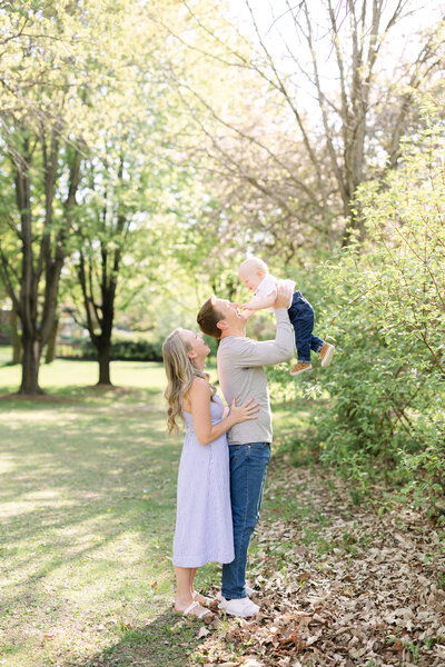on a sunny spring day, a father joyfully puts his baby into the air as the baby laughs and the mother stands behind dad, cheering him on
