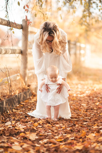 A stunning fall portrait in Kamloops of a mother and daughter with blonde hair. They are both wearing white dresses. Mom is standing and bent over to hold up her daughter while she stands. Captured by Brittany Danielle Photography