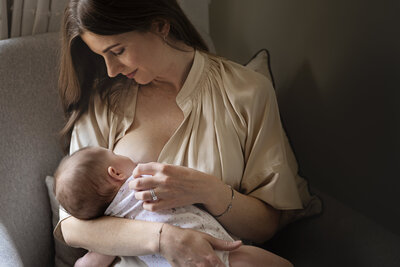A mother sits in soft natural light and breastfeeds her baby daughter during a relaxed at home photo shoot with London newborn photographer Jess Morgan