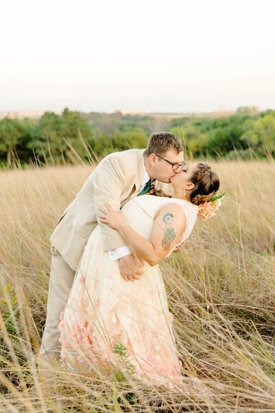 Bride and groom with their arms embracing each other as the groom is dipping the bride back while kissing her in a field at sunset during wedding portraits by Kansas City Wedding Photographer Sarah Riner Photography.