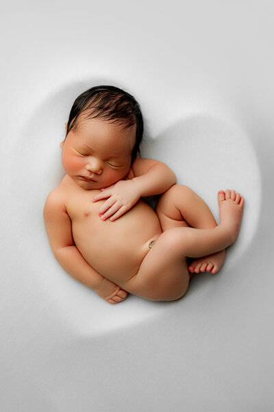 Newborn boy posed with hands holding head and toes peeking from behind his elbows on a brown backdrop