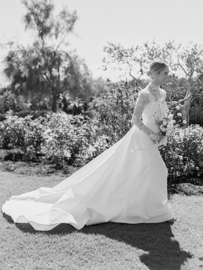 A bride and groom walk hand in hand down a grass aisle, smiling at their wedding guests as they clap. The wedding dress is body fitting, with full-length, semi-see-through sleeves.
