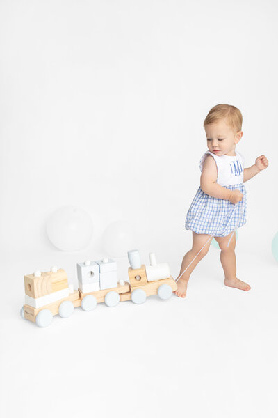 A one year old child in a traditionally smocked, blue gingham romper pulls a pastel wooden train behind them. A white studio backdrop is punctuated by soft white and turquoise balloons in the background.