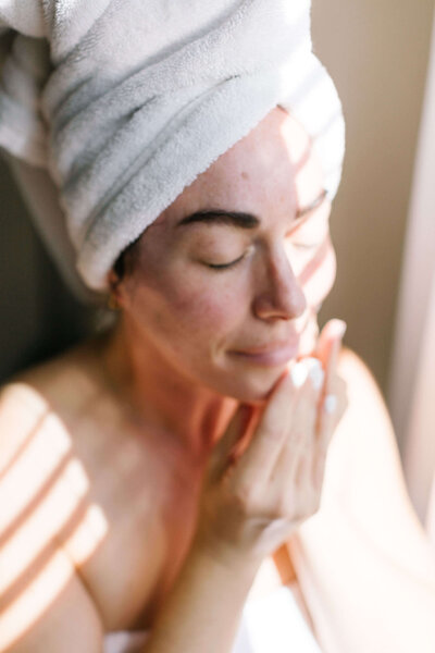 A woman in a towel  on her head stands by the window, enjoying the calmness of a sunny morning.
