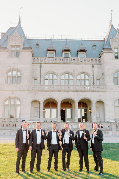 Ochre Court Newport | A group of six men in tuxedos joyfully pose with cigars in front of an elegant historic mansion.