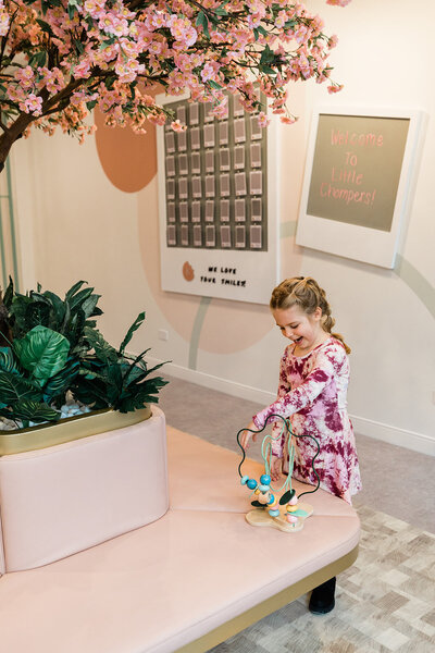 A happy, preschool-aged little girl in a floral dress, plays with a beautiful wooden toy in the waiting area of the best pediatric dentist near me. The waiting room features soft blush upholstered bench seating, accented with gold. In the background is a painted wall with geometric shapes in rust, blush, and soft sage green.