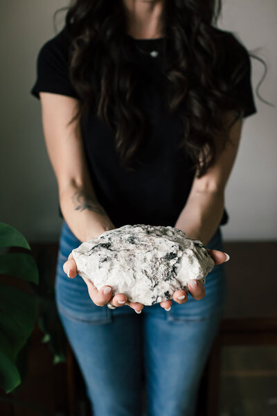 Moonstone founder Miranda holding a large raw moonstone crystal with soft hands and leafy greenery, symbolizing grounding, intuition, and spiritual brand identity.