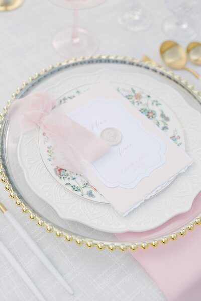 Elegant wedding reception details at Winbrooke in Brookshires, Massachusetts. The image features a beautifully set table with delicate pink napkins, floral china, and a vintage-inspired invitation. Felisha, a destination wedding photographer based in New England, captures timeless moments and beautiful details at weddings in Massachusetts, New England, and worldwide.