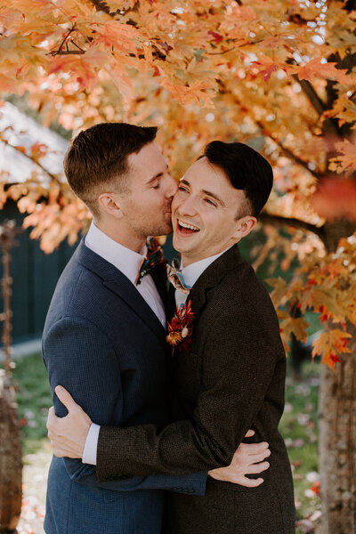 Two grooms hug under a tree with autumn colors