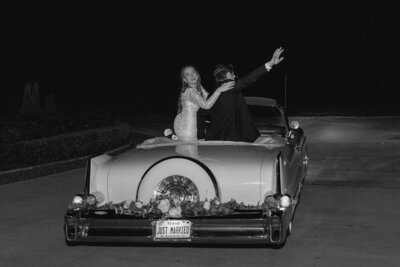 Black and white photo of bride and groom in their getaway car waving and surrounded by bubbles