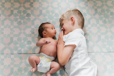 A baby and her older brother laying on the floor looking at each other cutely while being photographed by a Los Angeles family photographer.