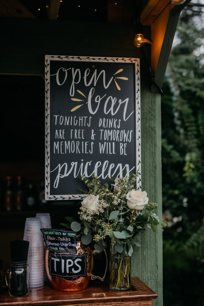 wedding bar decorative chalkboard sign that reads "open bar: tonights drinks are free and tomorrow's memories will be priceless." this sits above a bar with a tip jar.