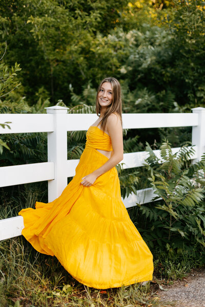 Senior girl in a long yellow dress twirling in front of greenery and a white fence