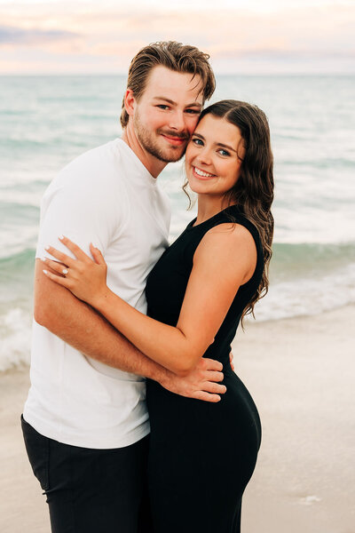 Woman with dark hair wearing black dress embracing man with white top in front of lake