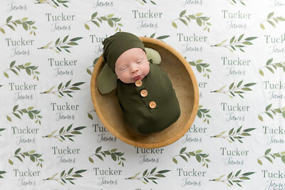 Newborn baby in a green wrap and hat in a small wooden bowl, on a blanket with his name on it.