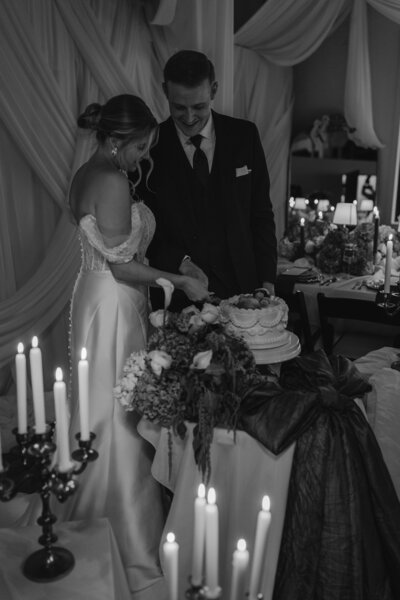 A bride and groom joyfully cut a tiered cake in a softly lit wedding setting. Elegant drapery and candlelight create a romantic, intimate atmosphere.