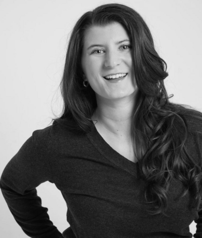 Black and white photo of woman with long, dark, curly hair wearing a black top with her hand on her hip and smiling.