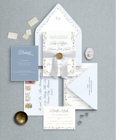 White Velvet ring box containing 3 rings, a men's watch, with greenery and white flowers, and wedding invitation suite in the background.