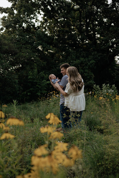 A family of three poses away from the camera in a field of yellow flowers. Mom and Dad are hugging while looking at their newborn baby.