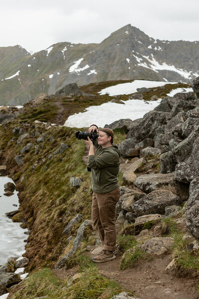 Sydney Breann, an elopement photographer, captures a couple in the Alaska mountains while standing on a rocky trail surrounded by snow patches and rugged peaks.