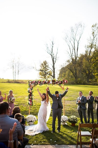 Bride and groom celebrating under a floral arch with guests cheering.
