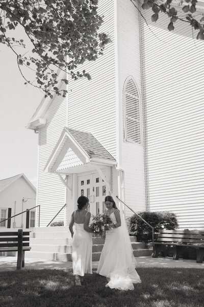 Women in wedding dress holding bouquet of flowers next to another women in white bridesmaid dress outside of a church