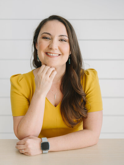 A woman in a yellow dress with her hand on her chin, smiling and looking at the camera during her brand photoshoot with Krista Marie