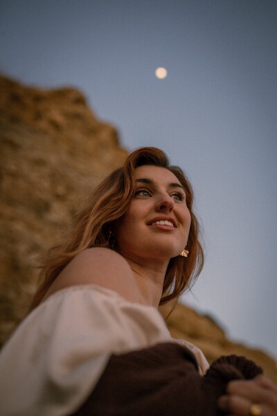A young woman with loose hair and off-the-shoulder blouse gazes upward, softly smiling beneath a twilight sky with the moon glowing above, framed by golden cliffs in the background.