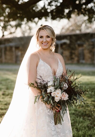 Bride in a white gown holding a bouquet, standing outdoors with sunlight highlighting her veil and background greenery. The scene captures the essence of modern bridal makeup trends, with expertly applied touches by an editorial bridal makeup artist enhancing her natural beauty.