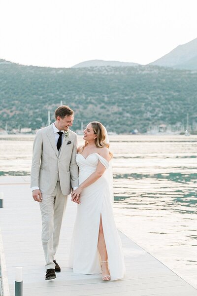 Bride and groom standing together on a dock at sunset, smiling against a mountain and sea backdrop during their intimate destination wedding