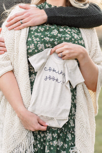 A close-up of a woman holding a "coming soon" onesie over her belly with her husband's arms embracing her from behind.