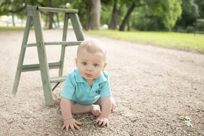 Adorable milestone session at Trinity Park, with the little one standing beside a charming mini ladder. The serene natural backdrop of trees and green space frames the moment, as the child reaches out with curiosity, dressed in a cute outfit and full of joy