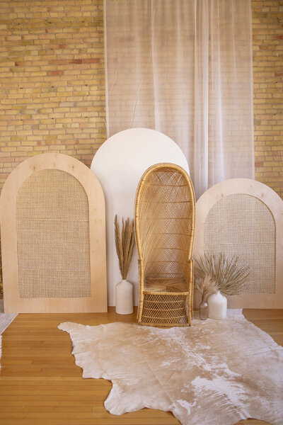 Two caned arches and one white wooden arch with a peacock chair in front. Pampas grass in vases on either side of the chair.