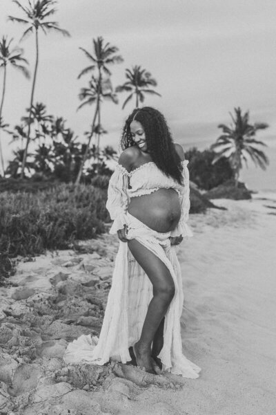 Black-and-white maternity photo of an expecting mother smiling on a Hawaiian beach surrounded by palm trees — elegant and emotive Oahu maternity photography by Leslie Carbajal Photography